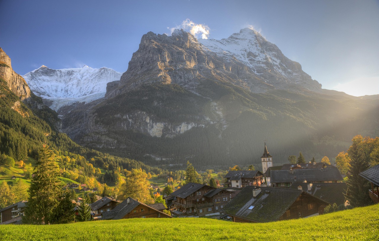 Traditional Swiss Alpine village with wooden chalets and church nestled in green valley, snow-capped mountain peaks and autumn trees under sunlit blue sky.