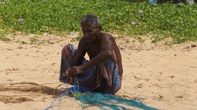 An elderly man on a sandy beach mending a fishing net