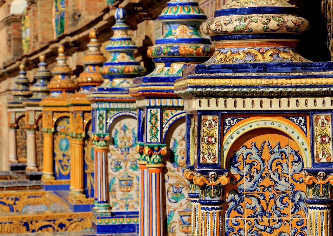 Siviglia a Giugno. Questa è una splendida immagine che mostra una serie di colonne decorate con ceramiche policrome in tipico stile spagnolo-andaluso, probabilmente parte della famosa Plaza de España di Siviglia.
Elementi principali:
Colonne ornate: Una fila di pilastri riccamente decorati si allinea in prospettiva, ognuno sormontato da guglie a forma di cupola
Ceramiche azulejos: Le superfici sono rivestite con piastrelle di ceramica dipinte a mano con motivi tradizionali spagnoli
Colori vivaci: Dominano il blu cobalto, il giallo oro, l'arancione, il verde e il bianco crema, creando un effetto cromatico brillante e festoso
Decorazioni:
Motivi geometrici e floreali
Arabeschi e volute barocche
Elementi ornamentali rinascimentali
Disegni simmetrici negli archi
Architettura: Gli elementi mostrano influenze moresche e rinascimentali tipiche dell'architettura andalusa, con archi a ferro di cavallo e dettagli intricati
L'immagine cattura la ricchezza artistica e l'artigianalità della tradizione ceramica spagnola, con una luce solare che esalta i colori brillanti delle decorazioni.