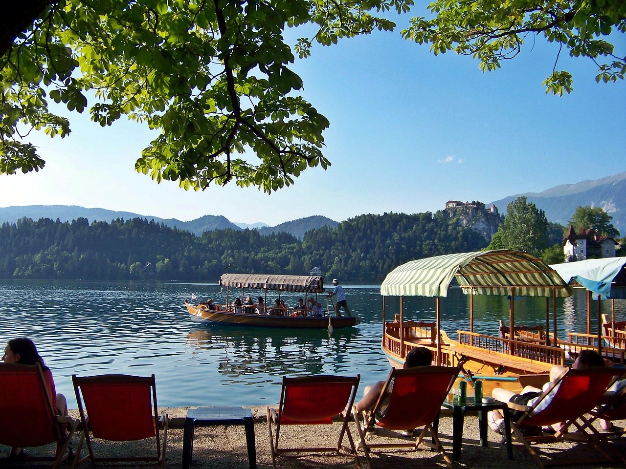 Lakeside scene with tourists relaxing in deck chairs under leafy tree, viewing traditional wooden pletna boats with striped awnings on tranquil blue water, with hilltop castle and forested mountains beyond.