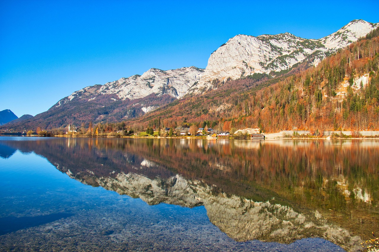 Serene Grundlsee lake with mirror-smooth water reflecting snow-capped mountains, autumn forest, lakeside buildings under vivid blue sky in Austrian Alps.