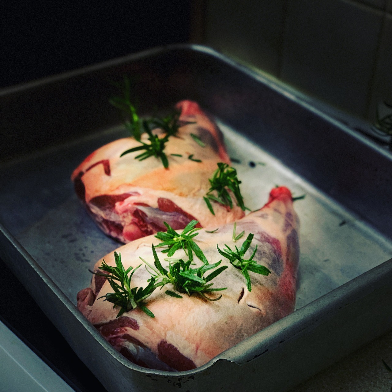 Two raw bone-in lamb joints topped with fresh rosemary sprigs in a metal roasting tin