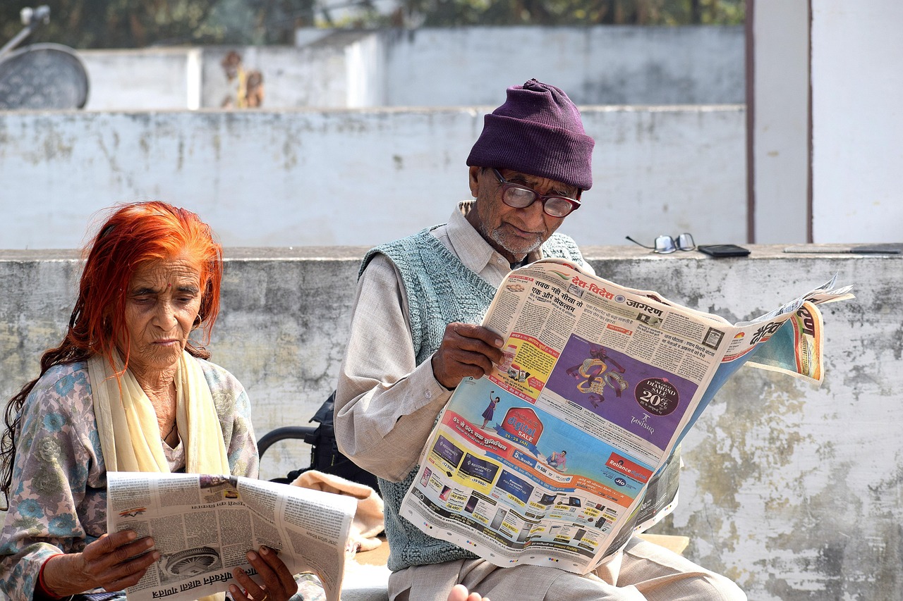 Older couple sitting together indoors reading a newspaper.