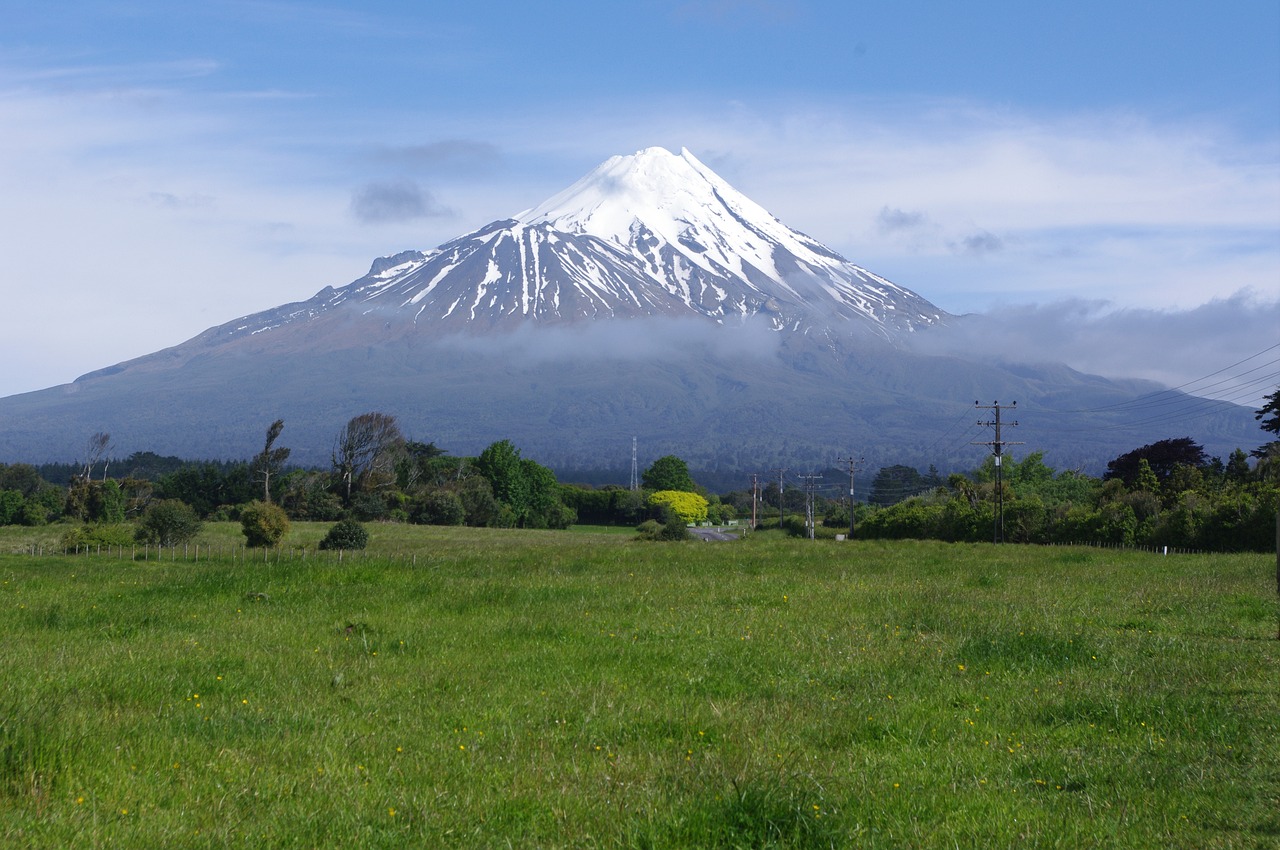 Majestic snow-capped Mount Taranaki volcano dominates the landscape above lush green farmland and rural countryside.