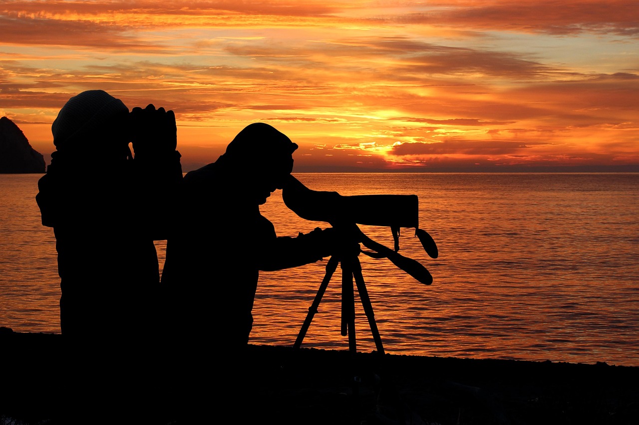 Pessoas usando um equipamento de observação apoiado em tripé diante do mar ao pôr do sol