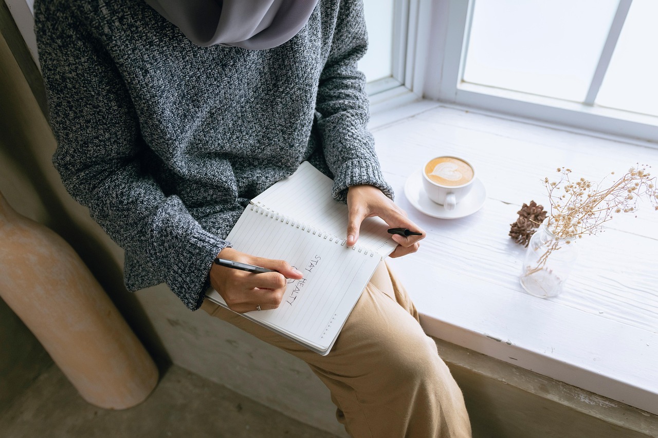 Mujer escribiendo en su diario junto a una ventana, con un café al lado, en un momento de calma y autocuidado