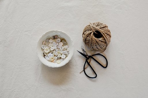 Bol de boutons récupérés, pelote de ficelle et petits ciseaux disposés sur un tissu neutre, matériel de loisirs créatifs prêt à réutiliser