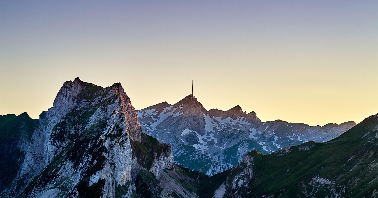 Dramatic alpine mountain landscape with rugged snow-capped peaks, deep valleys, layered ridges, and patches of green vegetation under clear blue sky.
