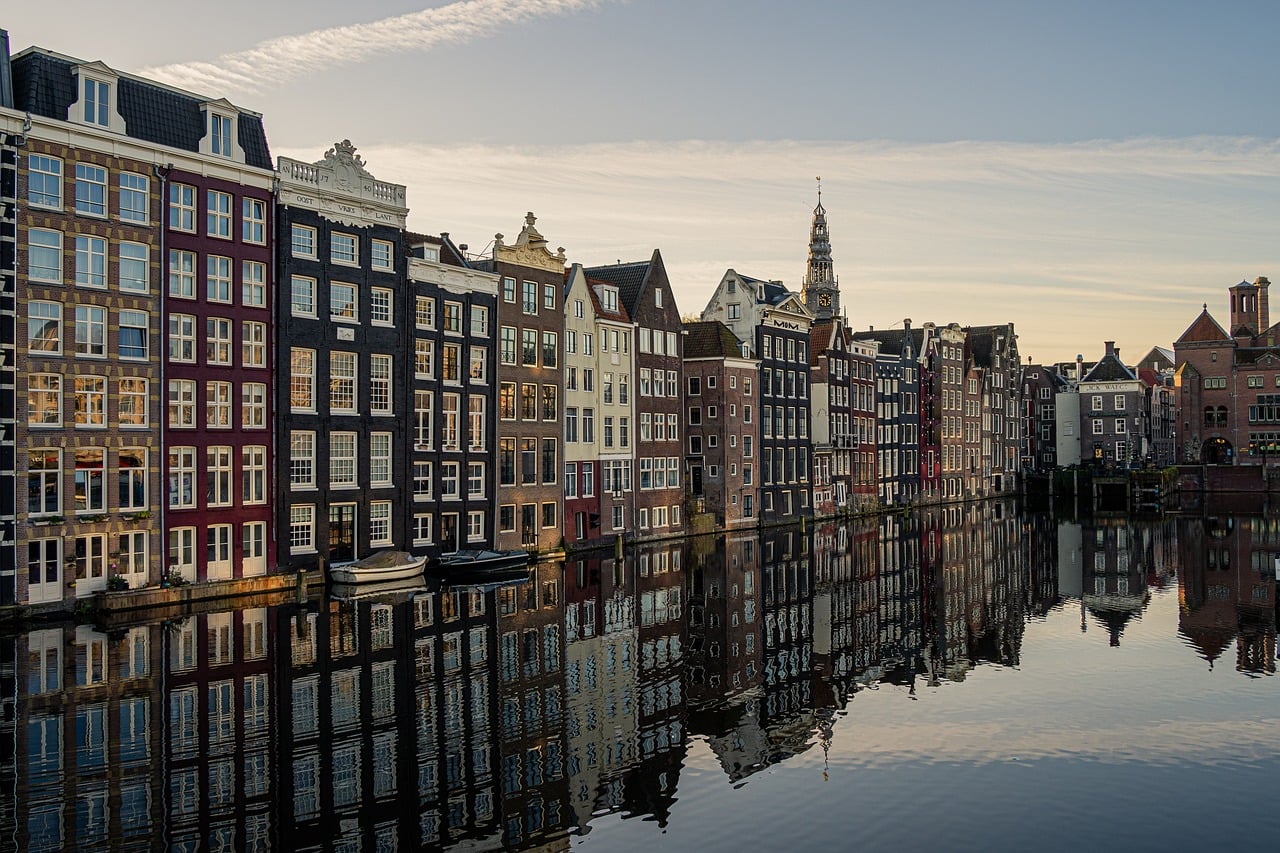 Traditional narrow canal houses in Amsterdam with colorful stepped gable facades perfectly reflected in still canal water, church tower in background, cloudy sky, iconic Dutch architecture.