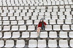 Random: empty seats, woman, independence, loneliness, hat, sunglasses, grey sweater, red coat, winter clothing, stadium, rows, outdoor, park, alone, sitting, steps, concrete, student, isolation, bench